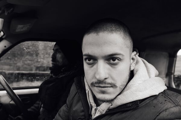 A black and white image of a young man looking out of a car window. An older man can be seen sat in the driver's seat. 