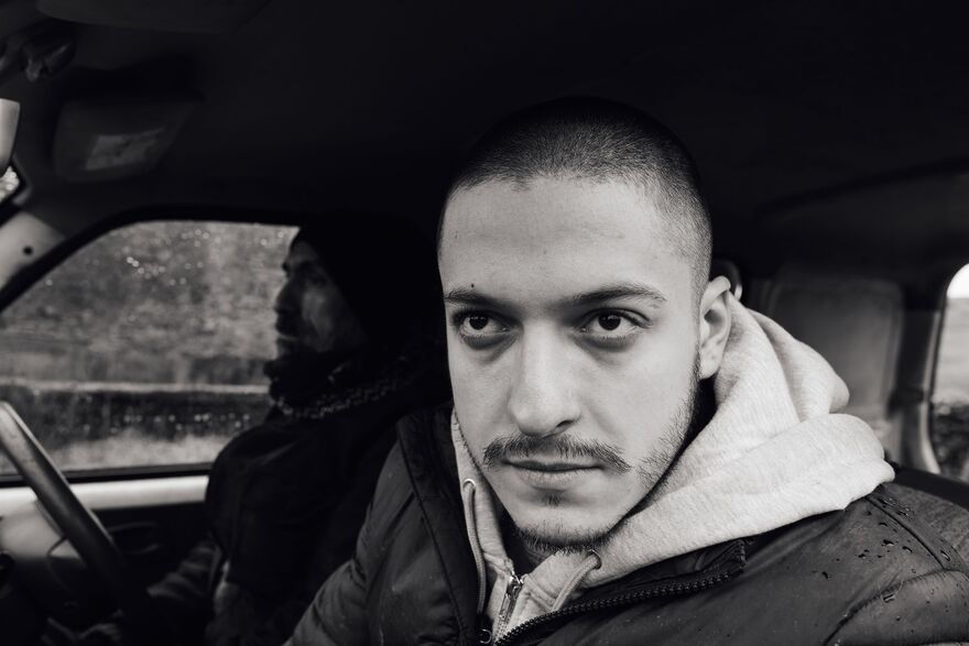 A black and white image of a young man looking out of a car window. An older man can be seen sat in the driver's seat. 