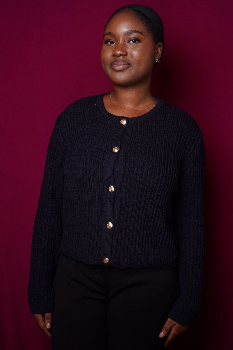 Zeinab wearing a black cardigan standing in front of a burgundy background