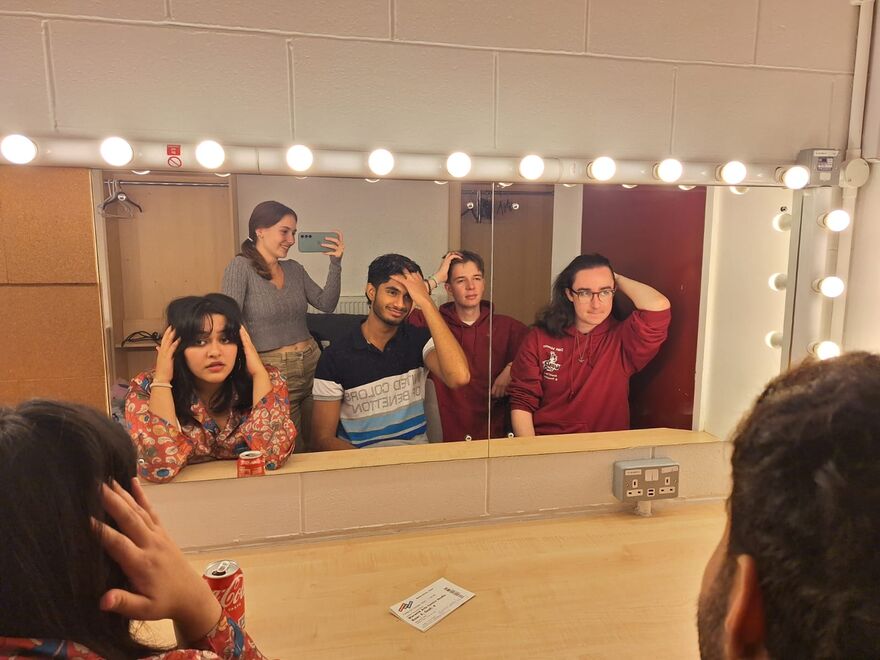A group of five people taking a mirror selfie in a dressing room, posing and adjusting their hair in front of a mirror.