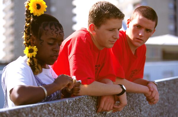 A group of three teenagers stand together leaning over a balcony. 