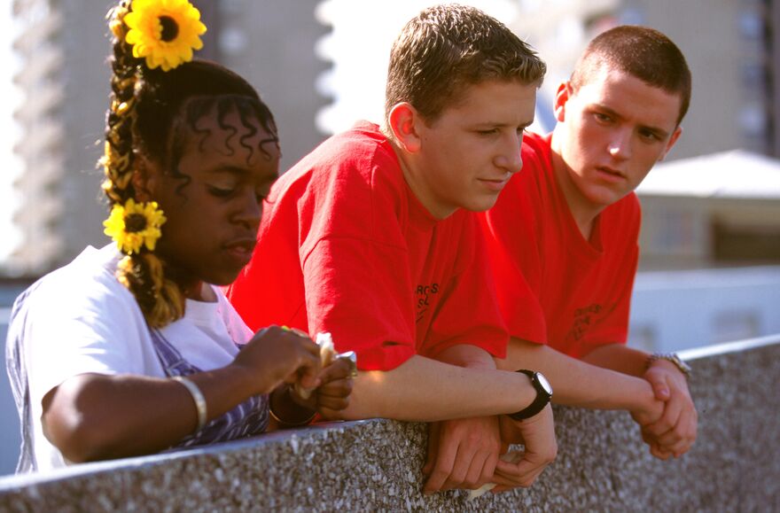 A group of three teenagers stand together leaning over a balcony. 