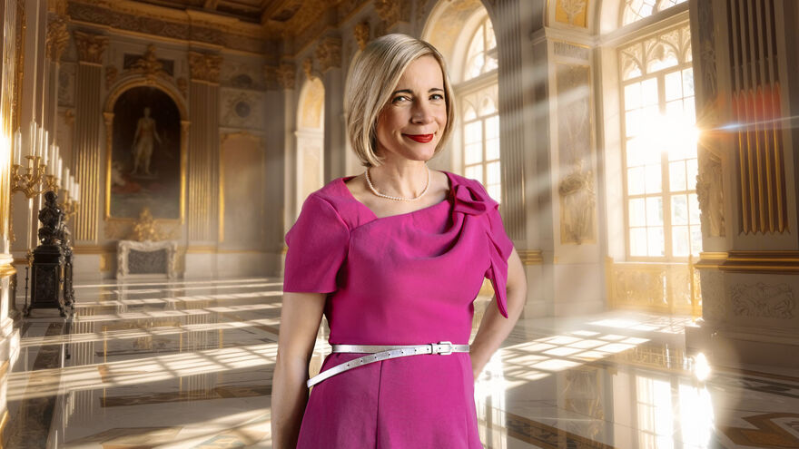 Lucy Worsley in a pink dress stood in a grand hall
