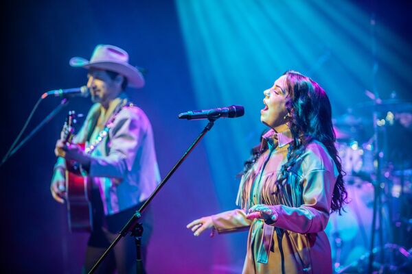 A woman singing into a microphone with a man in a cowboy hat and guitar signing into another microphone