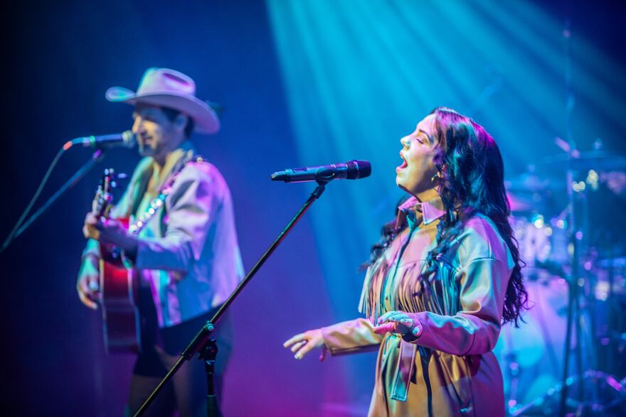 A woman singing into a microphone with a man in a cowboy hat and guitar signing into another microphone