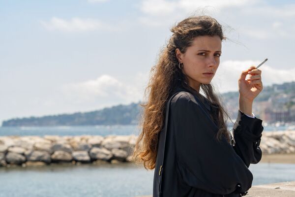 A young woman stands on a seafront. She is holding a cigarette, looking straight to camera. 
