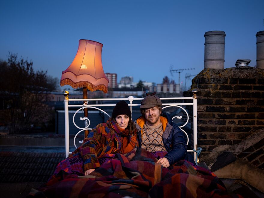 A couple sit up in a double bed on a roof top with a standing lamp behind them. They are dressed warmly.  