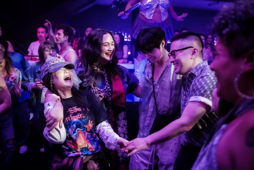 A group of four friends dance together at a club. They are bathed in purple light.