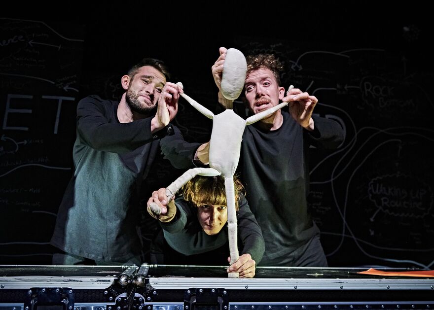 Fred, a two - foot - tall white cloth puppet , is stood dramatically with his arms and one leg stuck out , lit up against a darker background. Hi s three puppeteers , who control his different limbs , are wearing black and look ing expressive as they move Fred into position