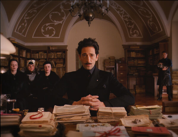 A man with a pencil moustache sits upright at a desk with his hands clasped. Three women stand in the background. 