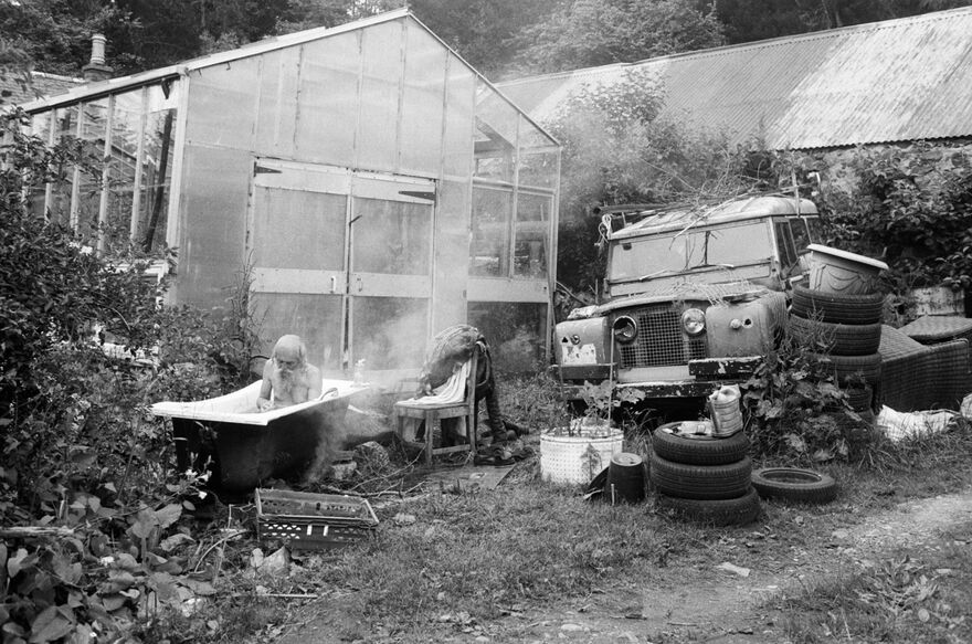 A black and white image of an old man sat in a bathtub outside. A green house, a car and stacks on tyres can be seen in the background. 