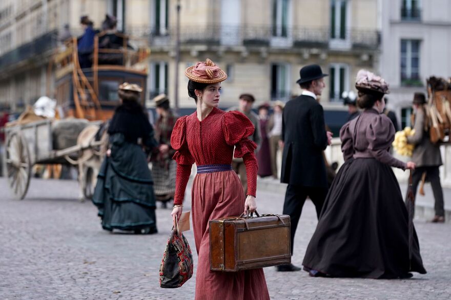 A woman in period dress looks behind her as she crosses a road. She is carrying luggage. 