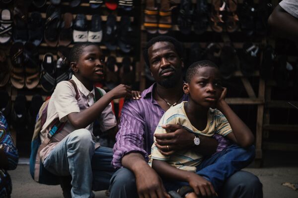 A father looks to camera as he holds his two boys close. They are sat in front of racks of shoes. 