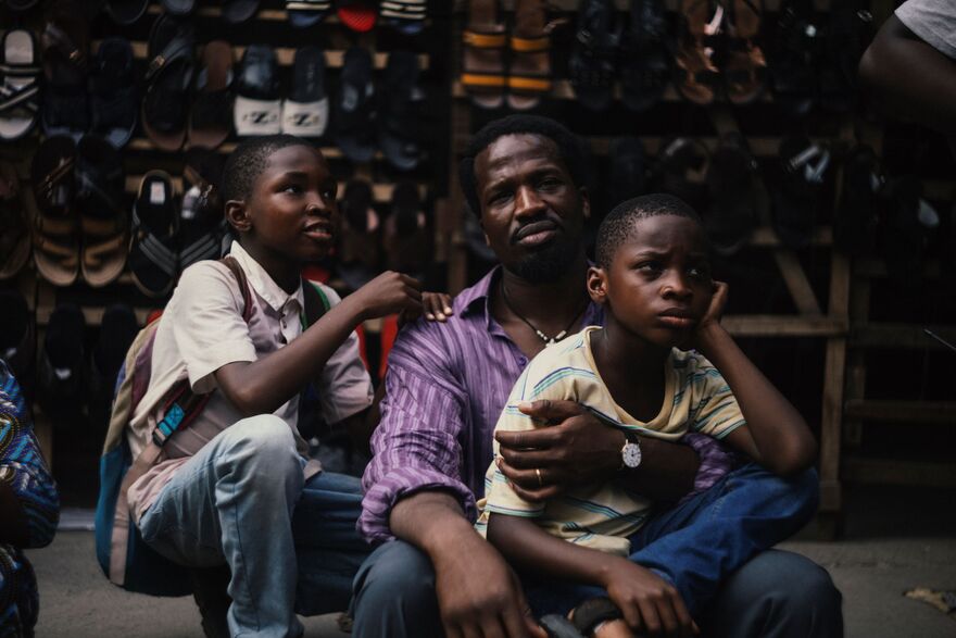 A father looks to camera as he holds his two boys close. They are sat in front of racks of shoes. 