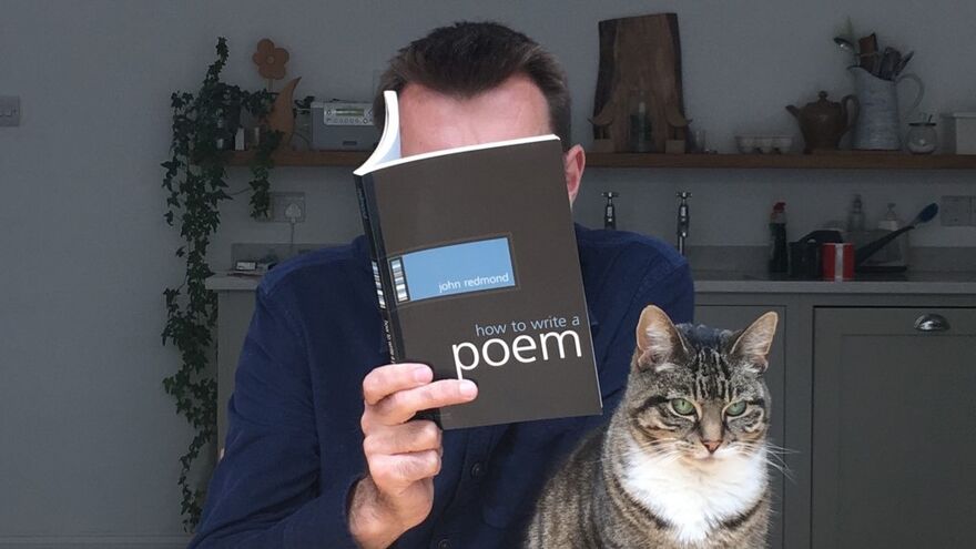 Brian Bilston hides his face behind a book titled 'How to Write a Poem', while a tabby cat sits on the table next to him.