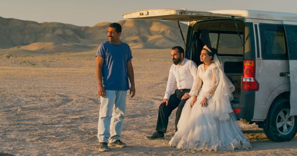 A bride and groom sit in the back of a white van with a man stood in front of them. They are in a desert landscape. A bride and groom sit in the back of a white van with a man stood in front of them. They are in a desert landscape.