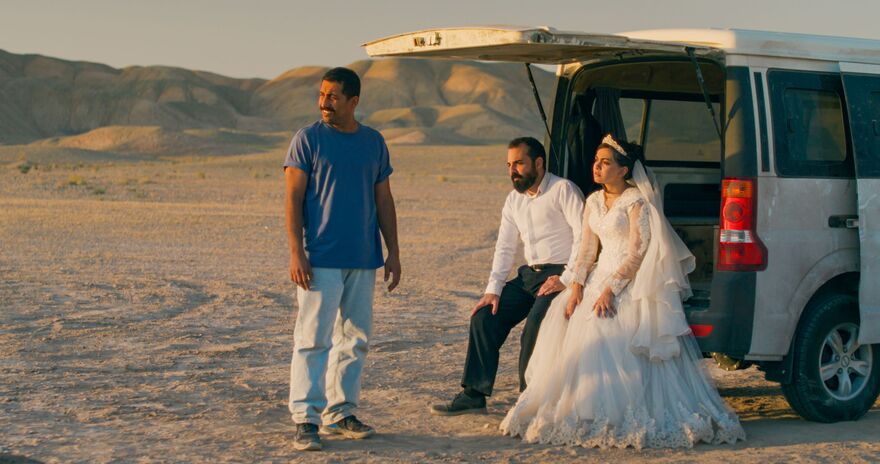 A bride and groom sit in the back of a white van with a man stood in front of them. They are in a desert landscape. 