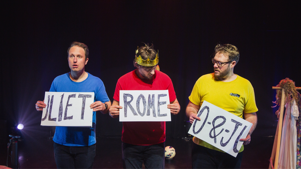 Three actors in brightly coloured t-shirts hold up jumbled signs spelling out 