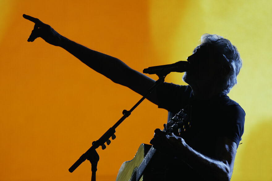 Roger Waters in silhouette holding a guitar singing into microphone with an orange background 