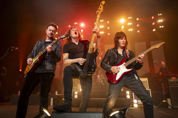 Three men play guitars at the front of a stage