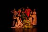 Nine dancers in colourful, flowing garments posed gracefully together in a tight cluster on a dark stage under warm lighting.