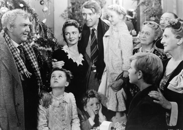 A black and white image. A family stand in front of a Christmas tree looking towards a man in a winter coat and scarf. 
