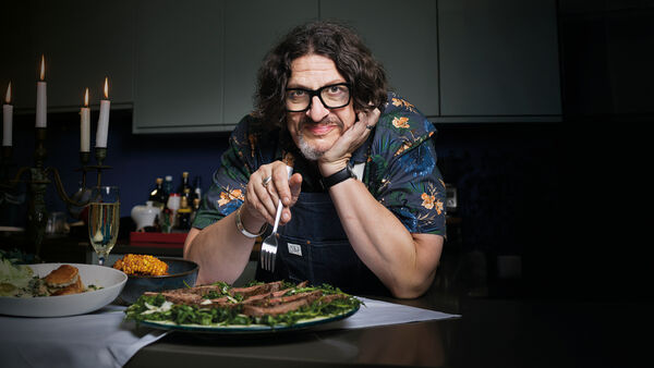 Jay Rayner leaning on a counter with fork in hand with a plate of steak and salad on the counter