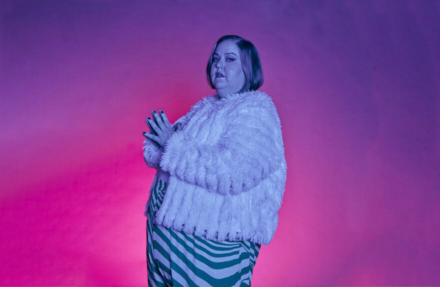 A woman looks to camera with her hands together. She is bathed in purple and pink light.