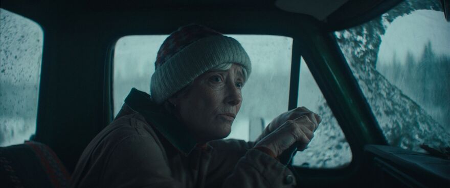A woman hunches over a car steering wheel. Snow can be seen on the windshield.