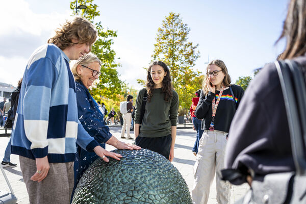 A group of people outdoors on a sunny day, with two of them touching and exploring a textured, dark green dome sculpture.