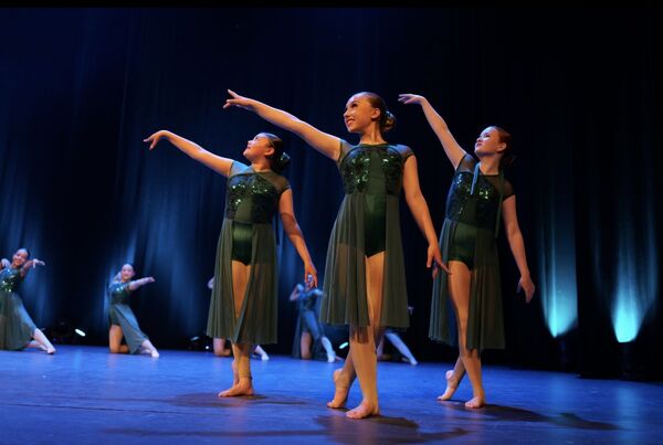 Dancers in dark green sequined costumes performing a graceful routine on a stage lit with blue lights.