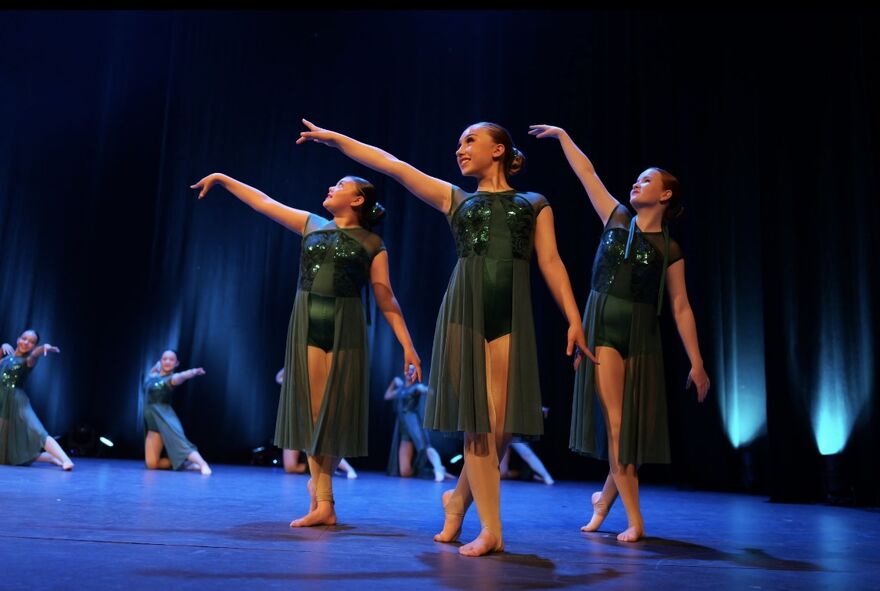Dancers in dark green sequined costumes performing a graceful routine on a stage lit with blue lights.