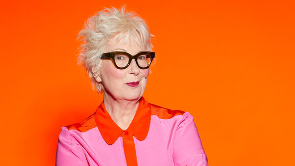 Jenny Eclair in thick framed glasses and a pink shirt with an orange trim, bright orange background