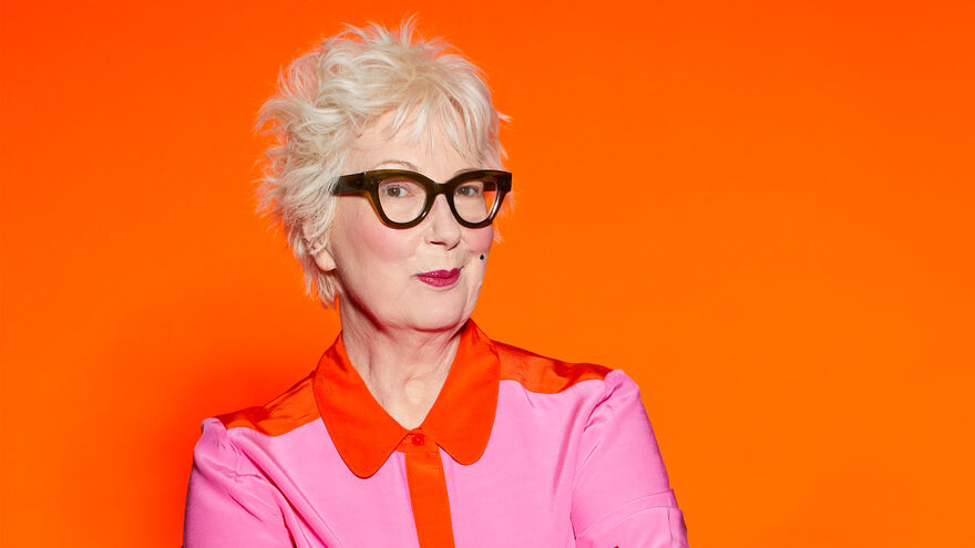 Jenny Eclair in thick framed glasses and a pink shirt with an orange trim, bright orange background