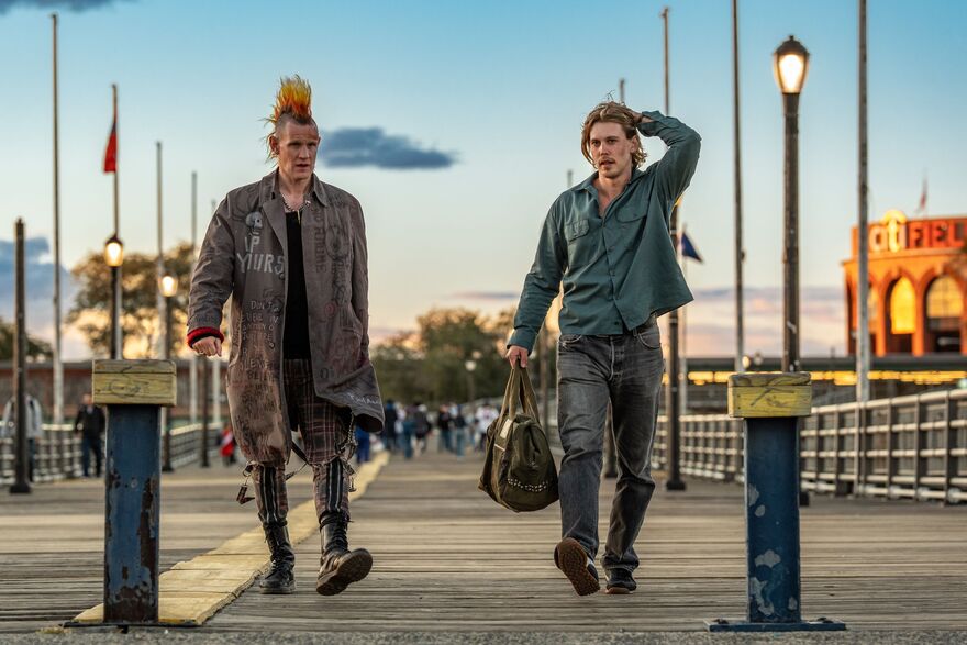 Two men walk together on a pier. One is dressed punk, with the other in denim. The man in denim is holding a duffel bag.