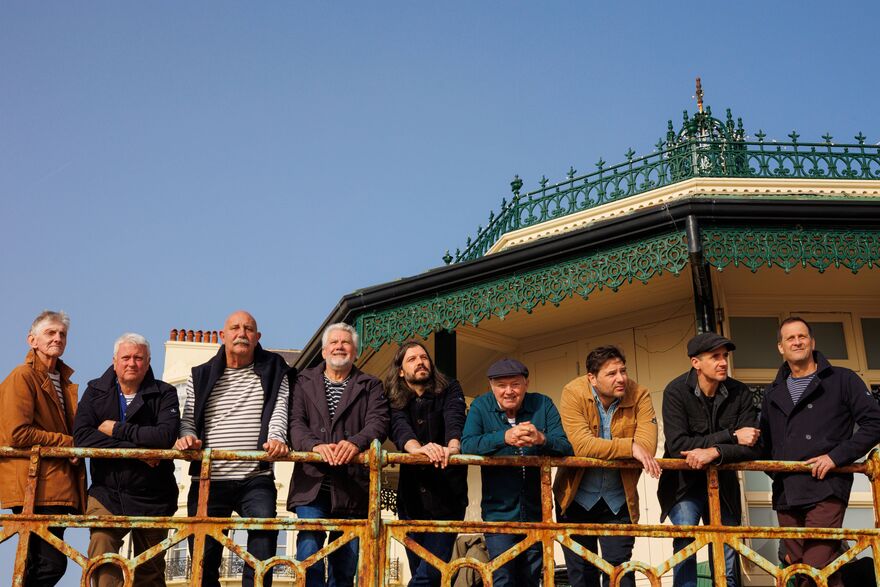The nine members of the folk group Fisherman's Friends standing together along a rustic railing in front of a seaside pavilion under a clear blue sky.