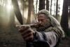 An older man with grey hair and beard aims a bow and arrow. He stands in woodland. 