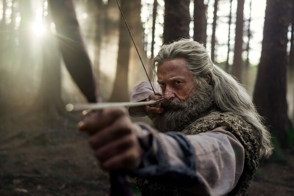 An older man with grey hair and beard aims a bow and arrow. He stands in woodland. 