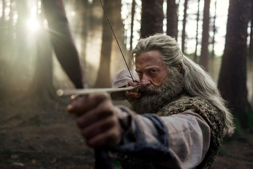 An older man with grey hair and beard aims a bow and arrow. He stands in woodland. 