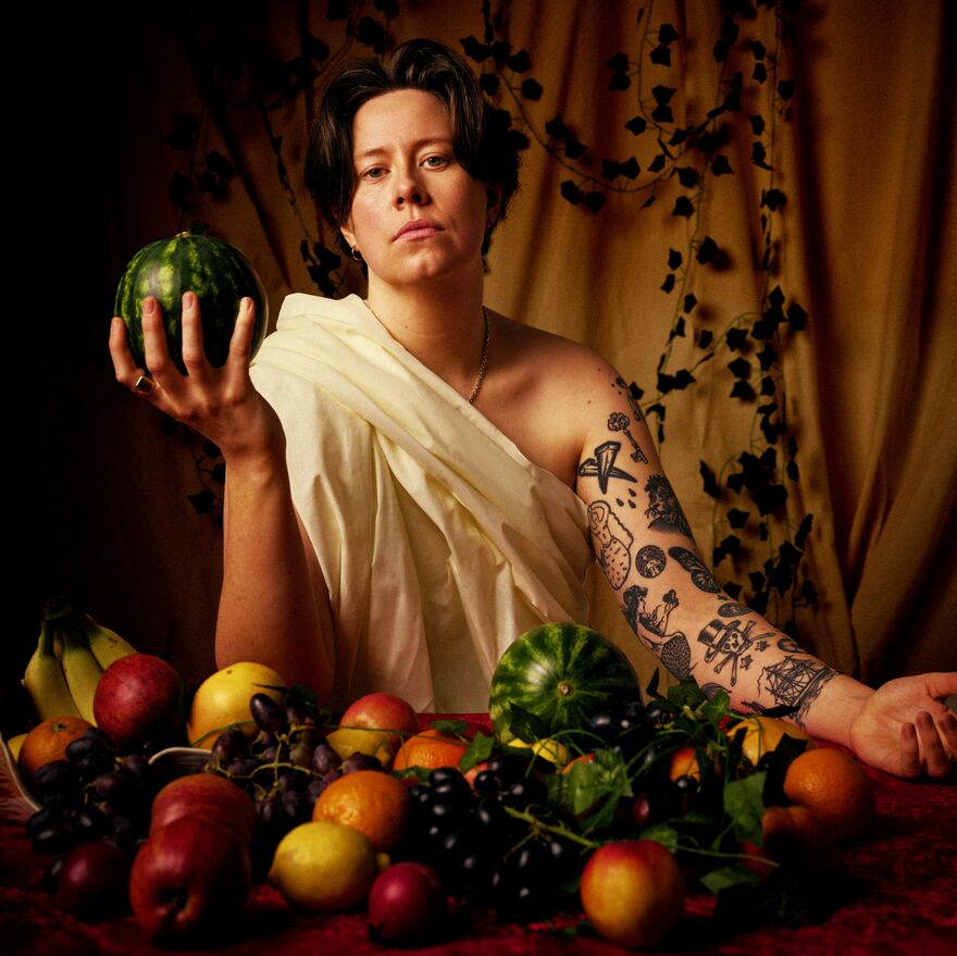 Chloe Petts in a white toga holding a melon with a selection of other fruits in front