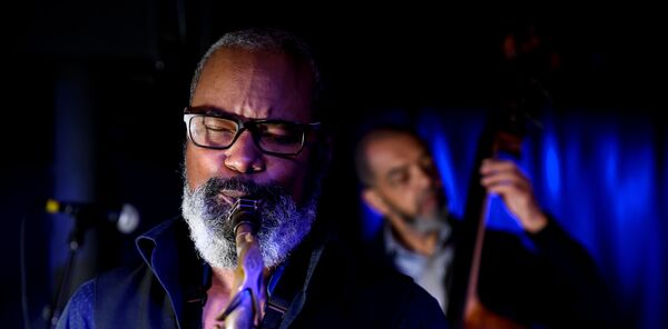 Saxophonist Denys Baptiste playing live with his eyes closed, while bassist Gary Crosby plays the double bass in the blurred blue-lit background.