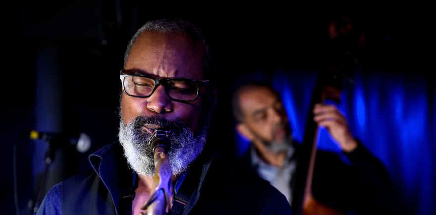 Saxophonist Denys Baptiste playing live with his eyes closed, while bassist Gary Crosby plays the double bass in the blurred blue-lit background.
