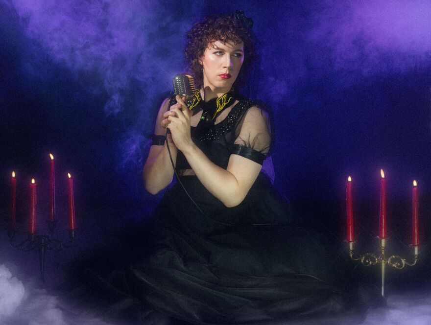 Sam Nicoresti in alon black dress holding a microphone surrounded by candles