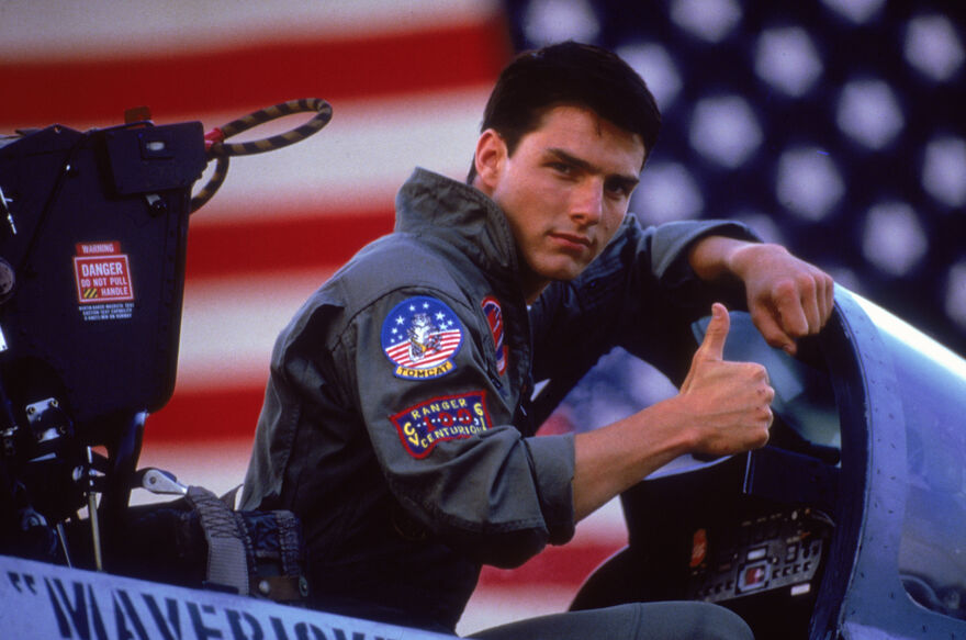 Tom Cruise as Maverick sitting in an aircraft with his thumb up.