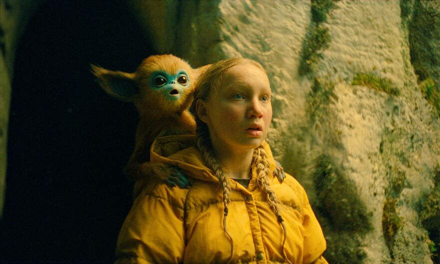 A young girl stands in front of a cave entrance with an ochi (small fantastical creature) on her shoulder. 