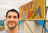 Creative workshop facilitator Benny Semp smiling in a selfie next to a colourful 'Crafty Kingdom' sign at an outdoor event.