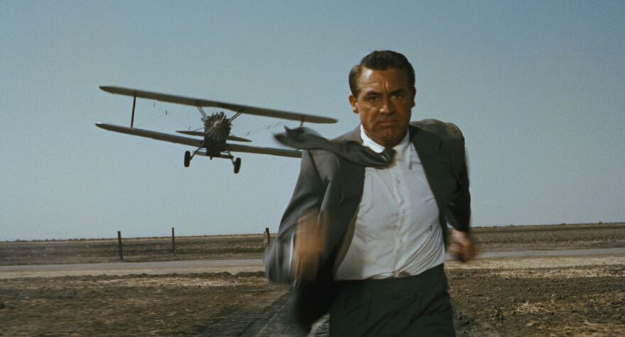 A man in a suit runs away from a low flying propeller plane on an air field. 