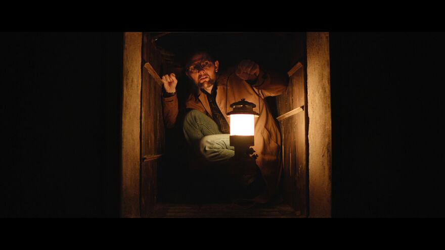 A man crouches in an small, enclosed space with a lantern. 