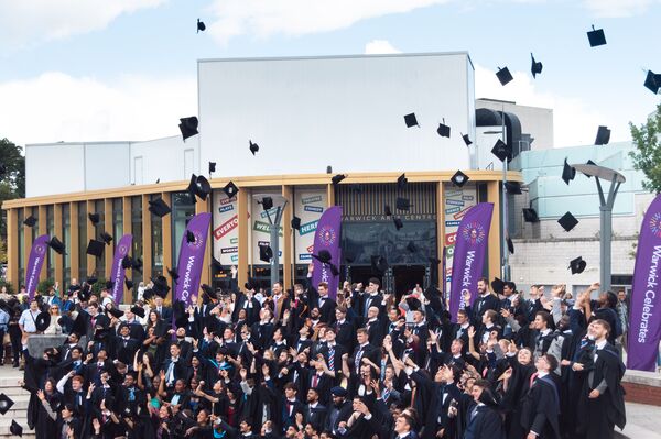 Graduands standing in a group throwing their caps in the air in front of Warwick Arts Centre