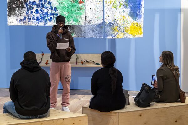 Three people sat on wooden boxes listening to a young man reading from paper into a microphone